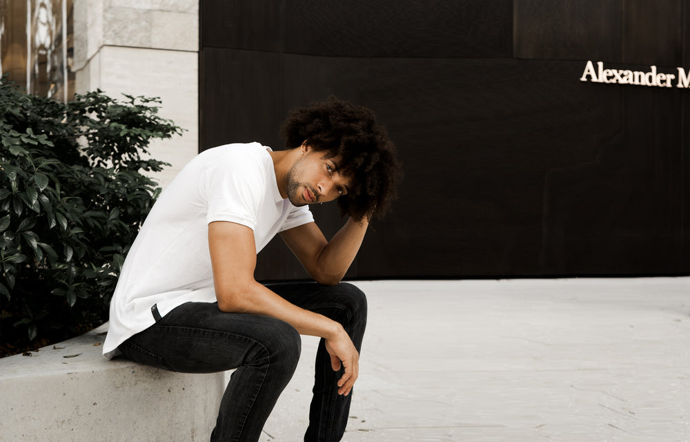 Light-skin, slim male model sitting outside of Brickell City Center in a white curved hem tshirt, slouched over picking curly-afro hair, while staring at the camera. 