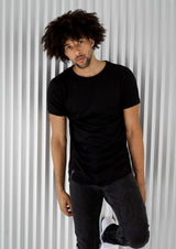Tall, light-skin curly-haired model wearing a premium men's black long-lined, fitted t-shirt leaning on a white wall.