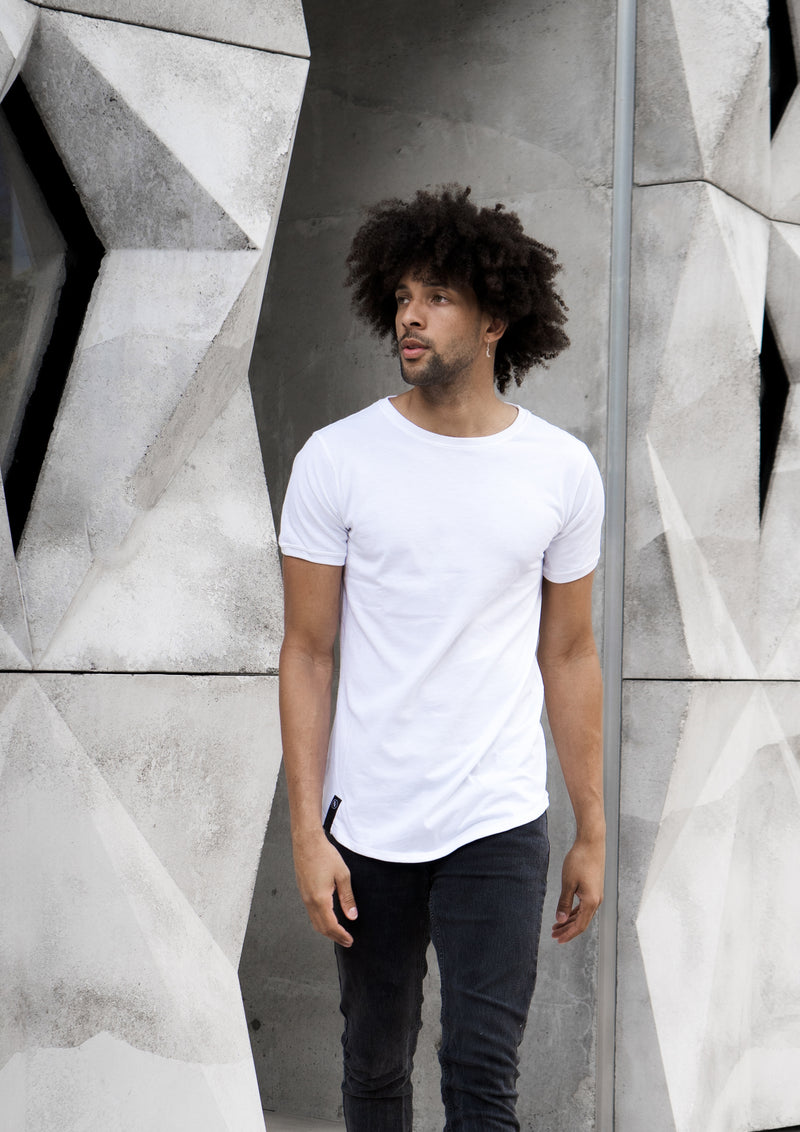 Candid photo of tall, light-skin curly-haired model wearing a premium men's white long-lined, fitted t-shirt walking away from rigid grey wall.